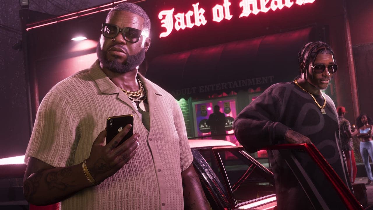 Boobie standing outside his club at night in sunglasses and gold chains, holding a phone, the neon Jack of Hearts sign above him.