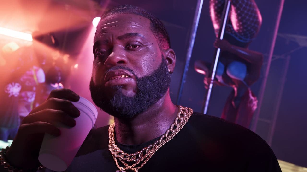Boobie in close-up inside his club under pink light, a drink in his hand and gold chains at his neck.