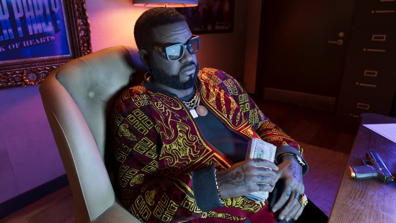 Boobie sitting back in an office armchair in a patterned jacket and sunglasses, holding a roll of banknotes.