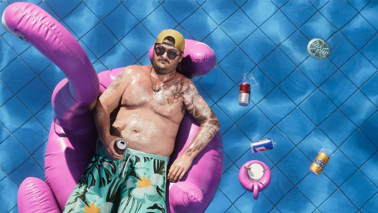 Cal photographed from directly above, floating on a pink flamingo ring in a swimming pool with empty cans drifting around him.