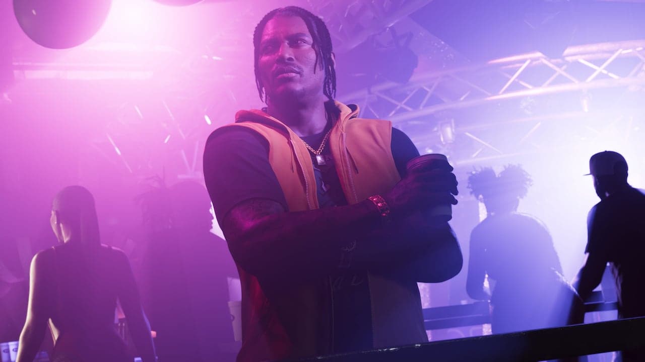 Dre'Quan standing with his arms folded on a club floor under pink and blue light, a cup in his hand.