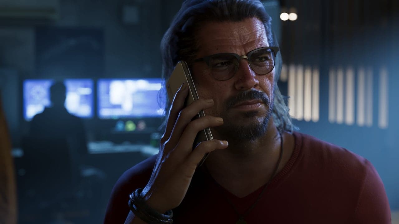 Raul taking a phone call in a dimly lit room, surveillance monitors glowing behind him.