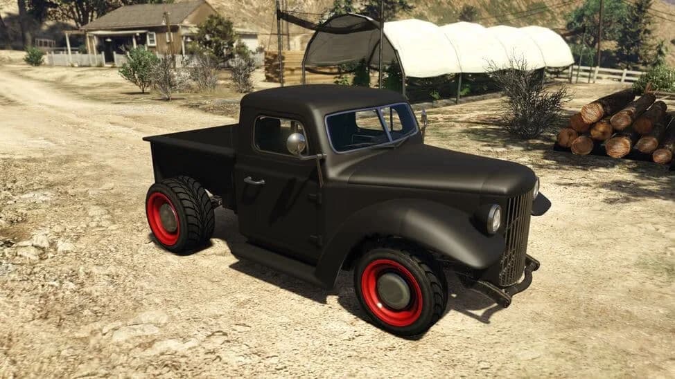 The Bravado Rat-Truck, a matte black 1940s-style hot rod pickup with red wheels, parked on a dirt yard, in GTA Online.