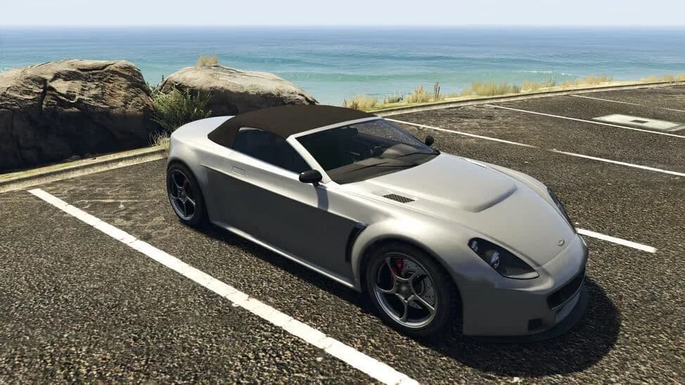 The Dewbauchee Rapid GT Cabrio, a silver modern convertible with its black roof raised, parked by the coast, in GTA Online.
