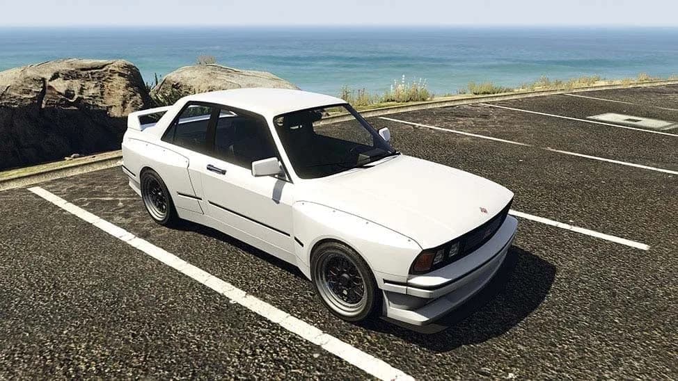 The Übermacht Sentinel Classic Widebody, a white 1980s saloon with flared arches and a rear spoiler, parked by the coast, in GTA Online.