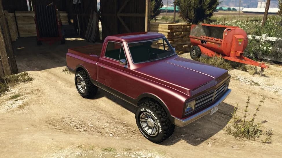 The Declasse Yosemite Rancher, a maroon lifted 1970s pickup on off-road tyres, parked by a barn, in GTA Online.