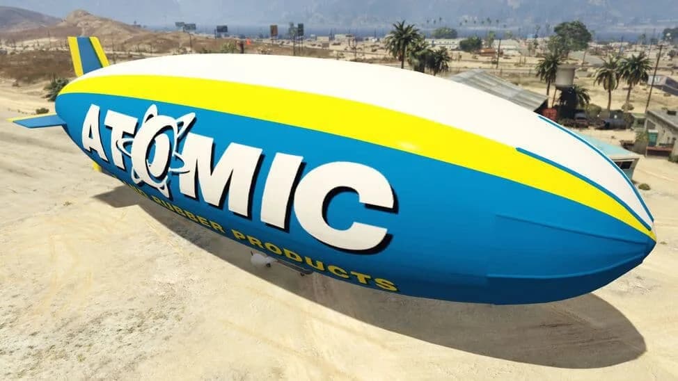 The Atomic Blimp, a blue, yellow and white airship in a tyre-brand livery, moored on a desert airfield, in GTA Online.